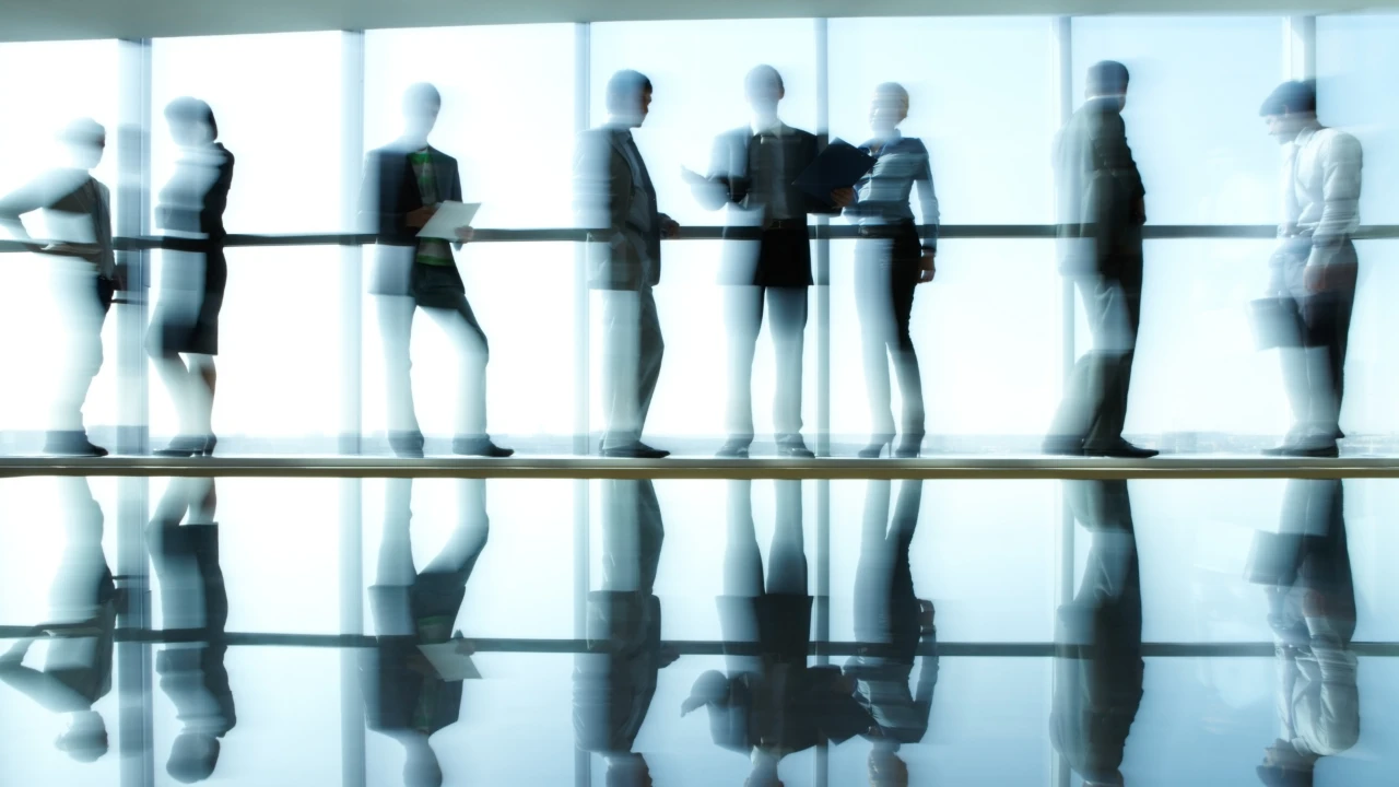 Silhouettes floues de professionnels en costume, alignés devant une baie vitrée lumineuse. Leurs reflets se mirent sur le sol poli, créant une ambiance moderne et éthérée.