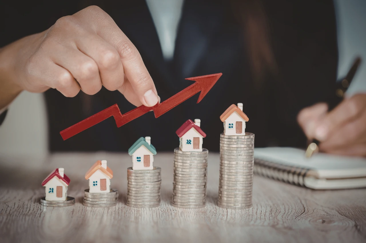 Main tenant une flèche rouge ascendante au-dessus de maisons miniatures sur des piles de pièces. Symbole d’investissement immobilier en croissance, avec carnet et stylo en arrière-plan.
