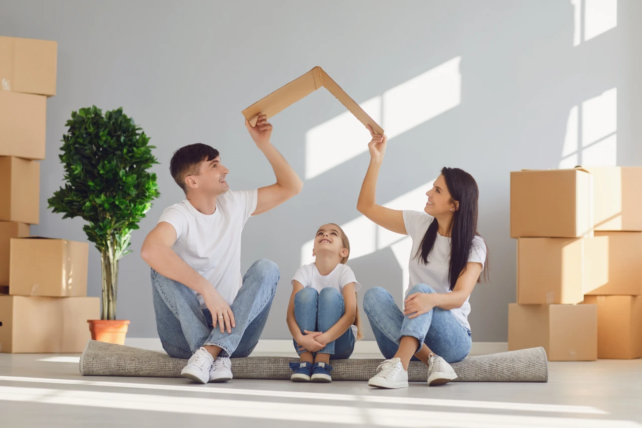 Famille assise sur un tapis dans une pièce vide, entourée de cartons. Parents tiennent un toit en carton au-dessus de leur fille souriante. Lumière naturelle, ambiance joyeuse et pleine d’espoir pour un nouveau départ.