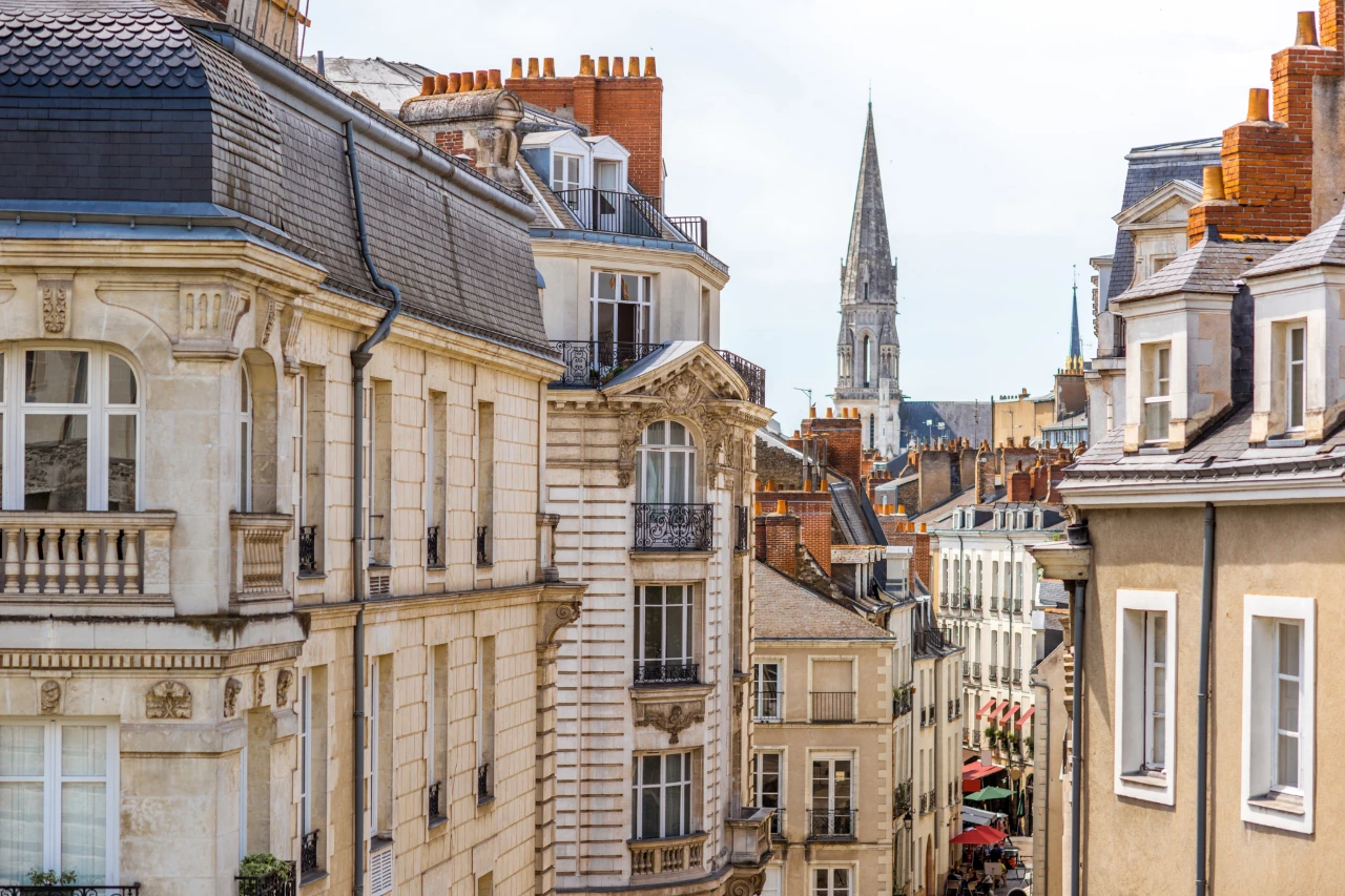 Rues étroites bordées d’immeubles haussmanniens aux toits d’ardoise et briques. Au loin, une flèche gothique domine le paysage urbain. Ambiance calme, lumière douce, typique d’une ville française historique comme Tours ou Orléans.